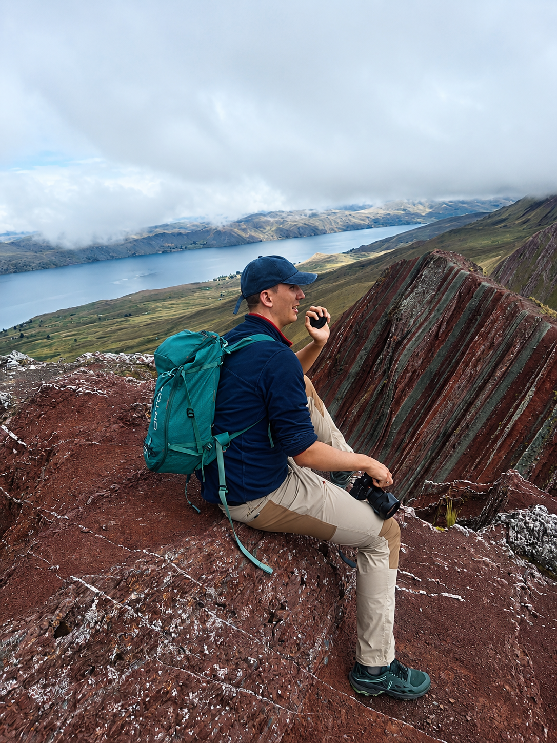 Excursionista con cámara descansa sobre roca roja con vista a laguna y montaña colorida en Pallay Punchu